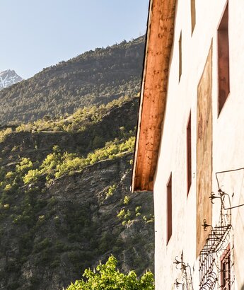 Wellness Angebot Vinschgau – Hotel Matill in Latsch Ein Teil eines Gebäudes mit einem Holzdach ist im Vordergrund zu sehen. Im Hintergrund erheben sich grüne Berge mit felsigen Abschnitten und schneebedeckten Gipfeln unter einem klaren blauen Himmel.